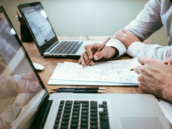 Two people sat next to each other between 2 laptops, writing notes together on a sheet of A4